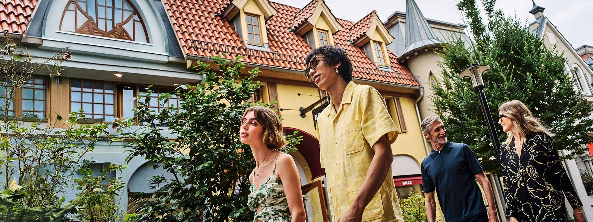 Four people walk outside in front of buildings with steep, red-tiled roofs and dormer windows on a cloudy day.