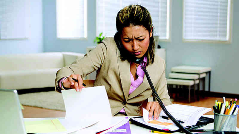 A businesswoman is sitting at a desk speaking on the phone with both hands sifting through two separate collection of notes.