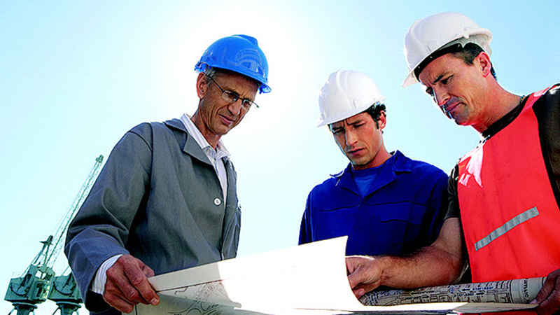 Three construction men are standing and viewing a set of blue prints with a large crane in the foreground.
