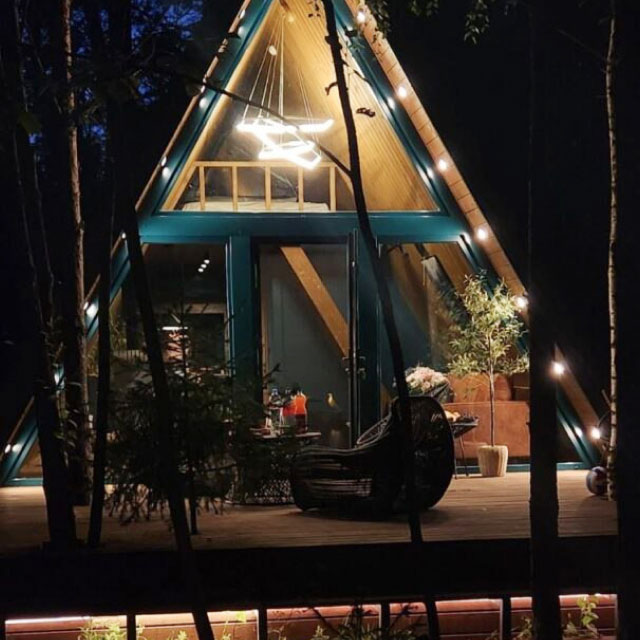 Домик с A-образной крышей ночью, уютно освещенный гирляндами вдоль деревянной внешней стены.