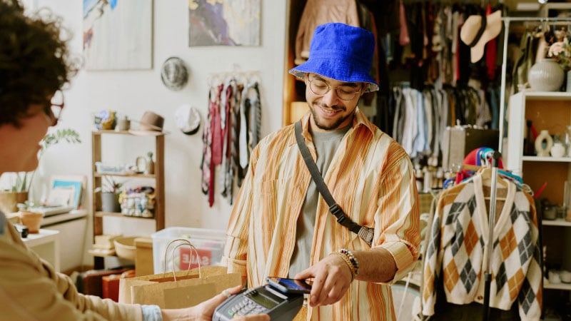 Человек в синей шляпе-котелке и полосатой рубашке улыбается, глядя вниз в уютной комнате, заполненной одеждой, сумками и декоративными предметами.