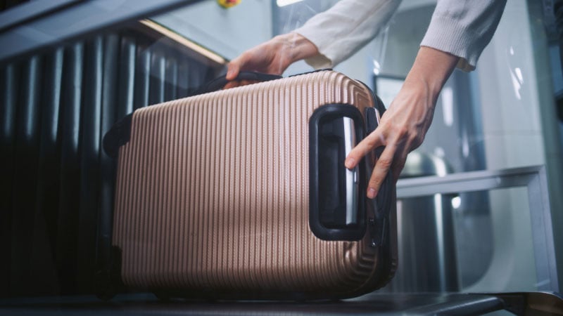 Руки поднимают ребристый бежевый чемодан, что намекает на путешествие или отъезд.