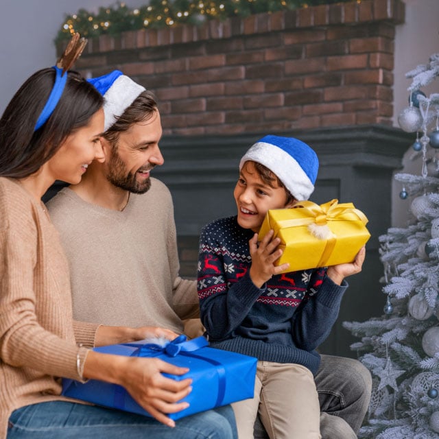 Веселая семья в праздничных шляпах сидит у украшенной елки и камина, обменивается ярко упакованными подарками и тепло улыбается.