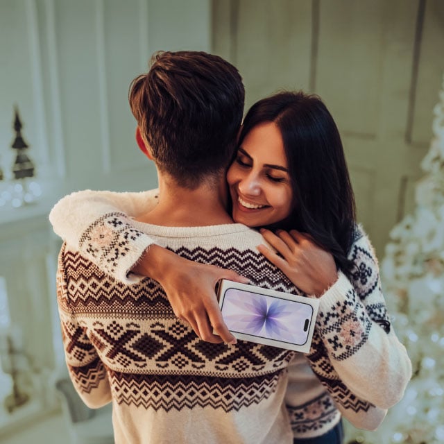 Женщина в уютном праздничном свитере обнимает мужчину, радостно улыбаясь. Она держит смартфон за его спиной.