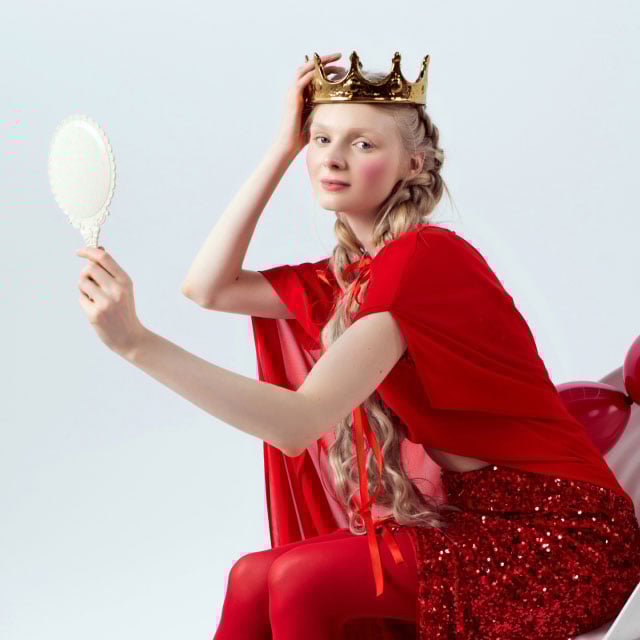 Женщина в красном платье сидит с изяществом, на голове у нее золотая корона, в руках она держит богато украшенное зеркало. 