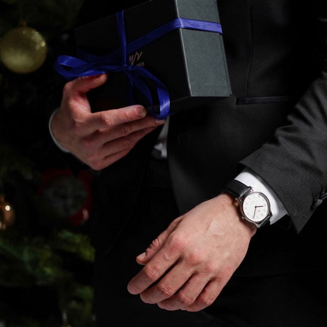 Мужчина в костюме держит подарочную коробку с голубой лентой, стоя рядом с украшенной рождественской елкой. На его запястье видны элегантные часы.