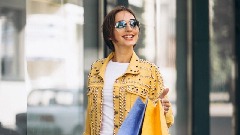 Улыбающаяся женщина в солнцезащитных очках и желтой куртке с заклепками держит в руках пакеты с покупками.