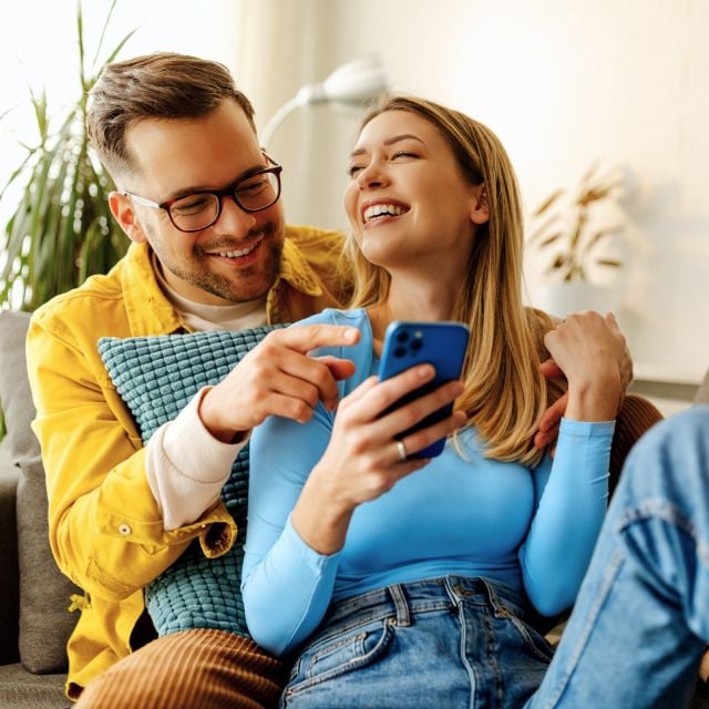 Радостная пара сидит на диване, увлеченно смотря в смартфон. Женщина в синем смеется, а мужчина в желтой рубашке держит подушку.
