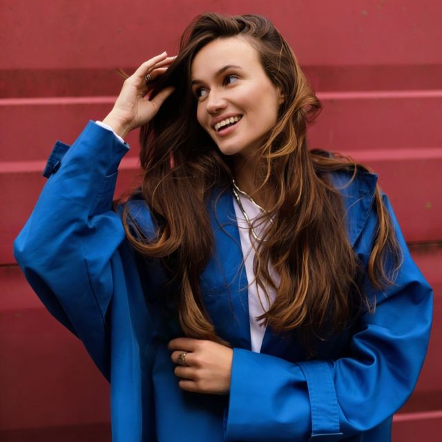 Улыбающаяся женщина в синем пальто стоит на фоне ярко-розовой стены. Ее длинные волосы ниспадают каскадом, она выглядит радостной и расслабленной.