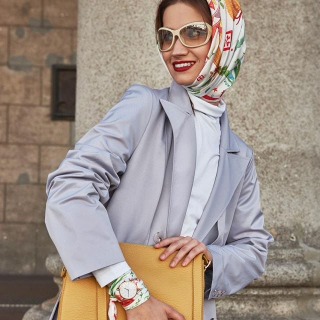 Стильная женщина в солнцезащитных очках и ярком платке уверенно улыбается. Она одела светло-серый пальто, держит в руках желтую сумочку и дополняет свой образ часами.