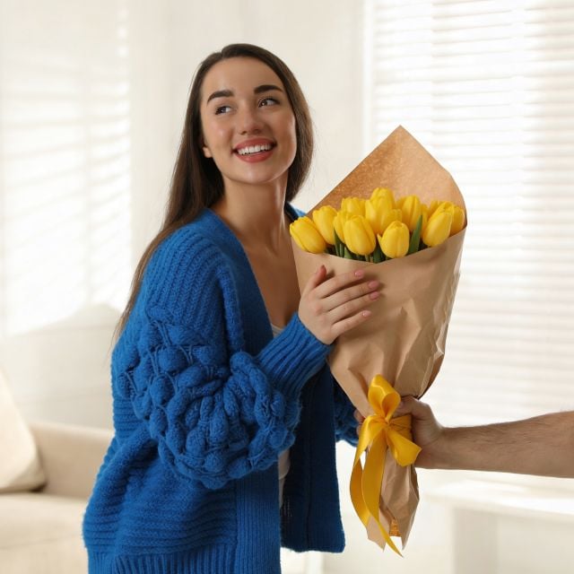 Улыбающаяся женщина в синем свитере получает букет желтых тюльпанов, завернутый в коричневую бумагу с желтым бантом.