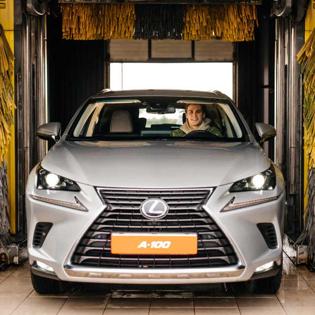 Серебристый внедорожник в автомойке, водитель улыбается . Ярко-желтые и черные моющие щетки окружают автомобиль.