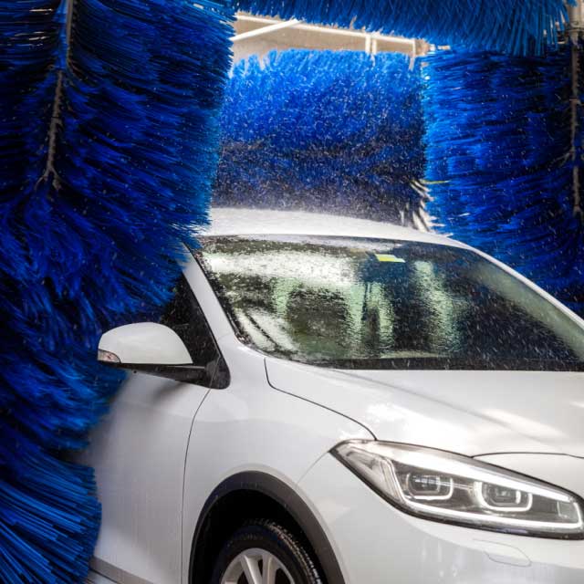 Белый автомобиль в автоматической автомойке, окруженный большими вращающимися синими щетками. Вода брызгает на автомобиль, создавая ощущение чистоты и свежести.