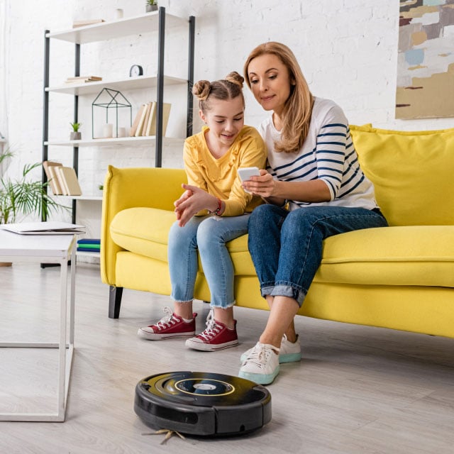 Женщина и девочка сидят на желтом диване, улыбаются и смотрят в смартфон. Робот-пылесос убирает деревянный пол в современной гостиной.