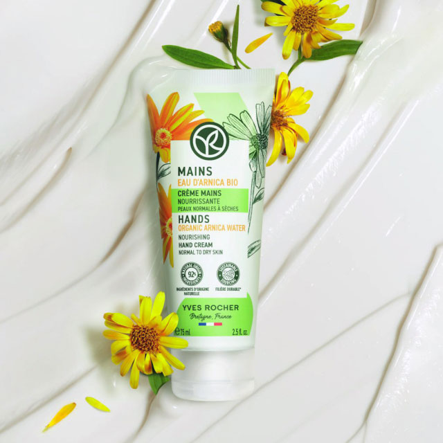 Тюбик крема для рук Yves Rocher с органической водой арники, окруженный желтыми цветами. Кремовая текстура на заднем плане, передающая питательность.