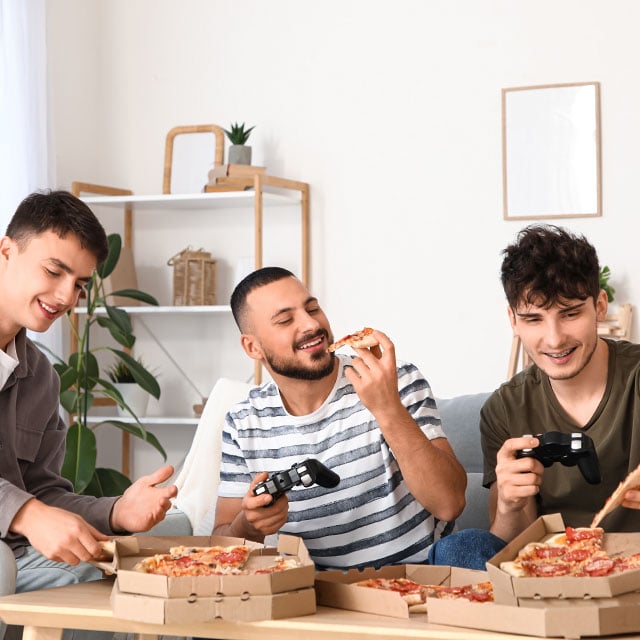 Трое молодых людей сидят на диване, едят пиццу и держат в руках игровые контроллеры, а на столе перед ними стоит несколько открытых коробок с пиццей.