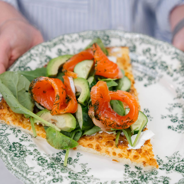 Кусочек хрустящей пиццы с добавлением свежего шпината, нарезанного огурца и копченого лосося на винтажной тарелке с цветочным узором, излучающей свежесть и изысканность.