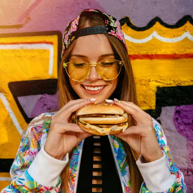 Улыбающаяся женщина в цветочной кепке и желтых очках держит гамбургер на фоне красочной стены с граффити. Ее выражение лица передает радость и удовлетворение.