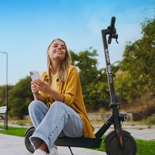Женщина в желтой кофте, улыбается и держит смартфон, сидит на черном электросамокате в парке. Небо ясное, на заднем плане видны деревья.