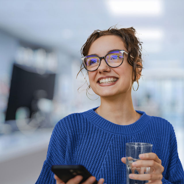 Улыбающаяся женщина в очках в синем свитере держит смартфон и стакан с водой.