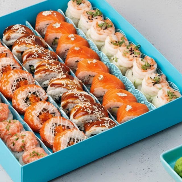 Бирюзовая коробка содержит ассорти суши-роллов, аккуратно выложенных рядами. Суши украшены ярко-оранжевым лососем, креветками и блестящим соусом.