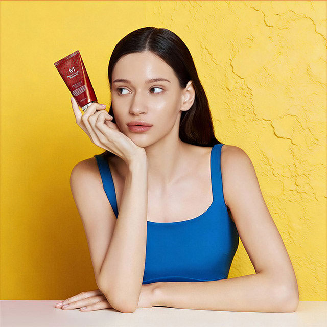 Женщина в синей майке держит красную тубу с косметикой, смотря в сторону на фоне текстурированной желтой стены.