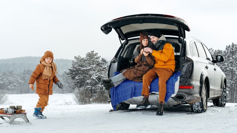 Семья, тепло одетая для зимы, сидит в открытом багажнике автомобиля, окруженном заснеженными деревьями. Ребенок, тянущий сани, подходит, улыбаясь.