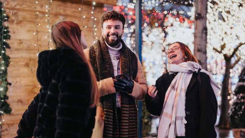 Группа из трех человек в зимней одежде весело смеется и болтает на улице, окруженная праздничными огнями.