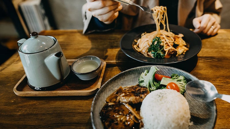 A person eats noodles with vegetables, with a plate of rice, hamburger patties, and vegetables, and a teapot with a small cup on a wooden table.