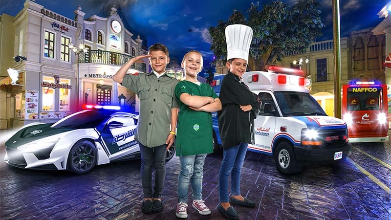 Three children dressed as a police officer, chef, and paramedic stand smiling in front of a police car, ambulance, and fire truck in a city-themed setting.