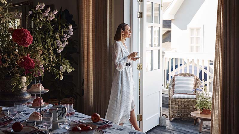 A woman in a white dress stands at an open door, holding a cup and looking outside; a dining table with flowers and food is set nearby.